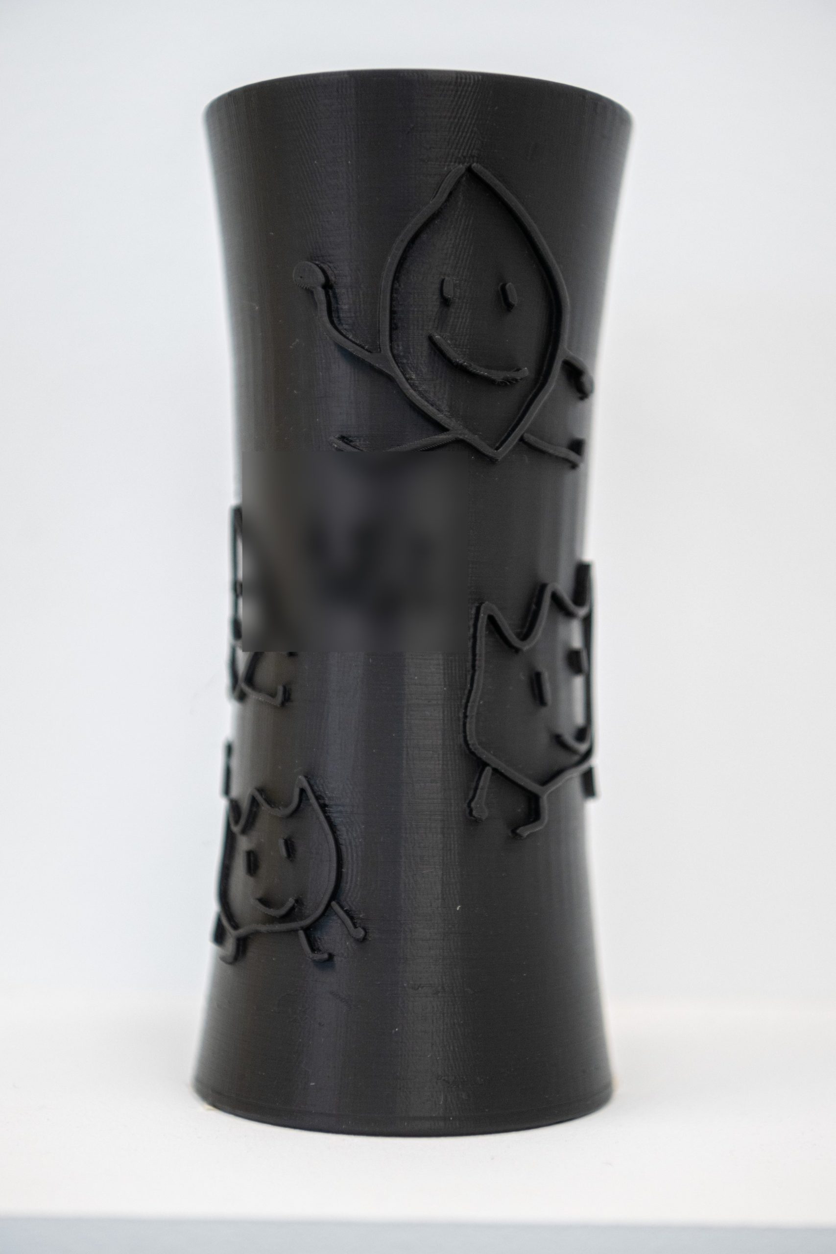Black 3D-printed vase carrying raised outlines of children's drawings — smiling round figures in relief (a handwritten name is obscured)